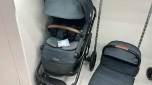 Two baby strollers in a store aisle, dark blue fabric and brown handles, one upright with a tag and seat cushions, another folded nearby.