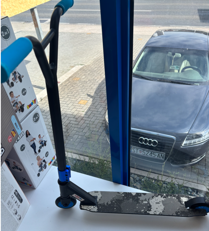 Slika zaslona 2026-07-09 u 12.38.22 Blue children's kick scooter on a stand inside a shop, seen through a bus-like window with a car parked outside.