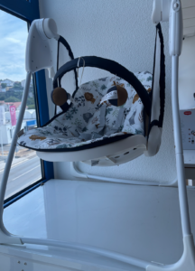Infant baby swing seat hanging from a white frame inside a store, with floral-pattern fabric and safety harness in the seat.
