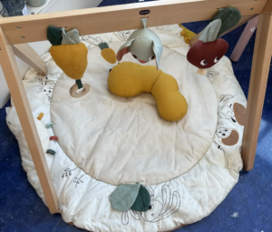 Wooden baby activity gym with a circular padded mat and hanging plush fruits: an orange carrot, a yellow pear, and a red apple over the center cushion.