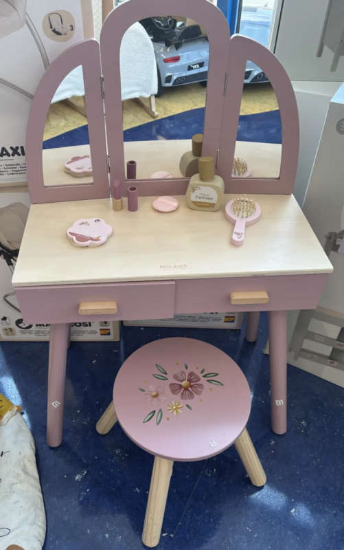 Pink children's vanity with three mirrors, small stool, and pretend makeup bottles on a wooden desk surface