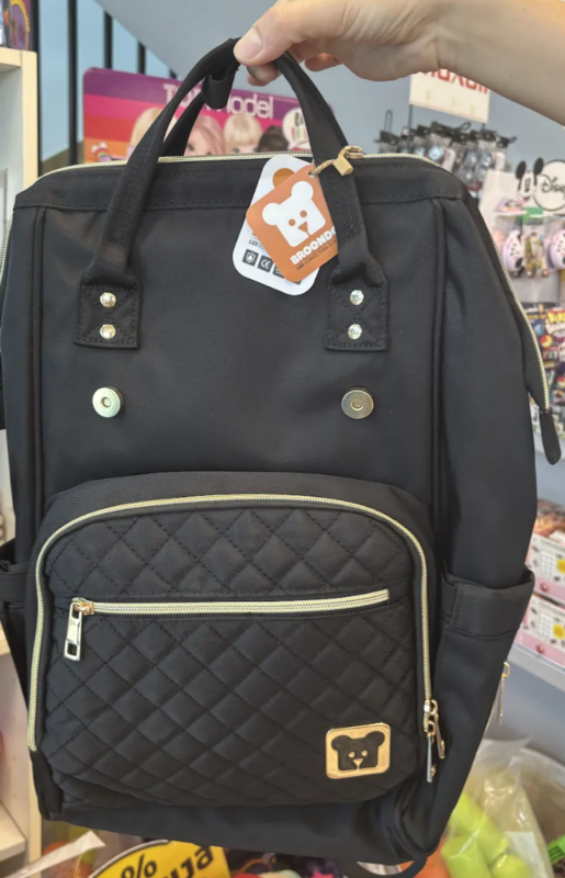 Black quilted backpack with a front zip pocket and gold-tone hardware on display in a store, tag with a bear logo hanging from the handle.