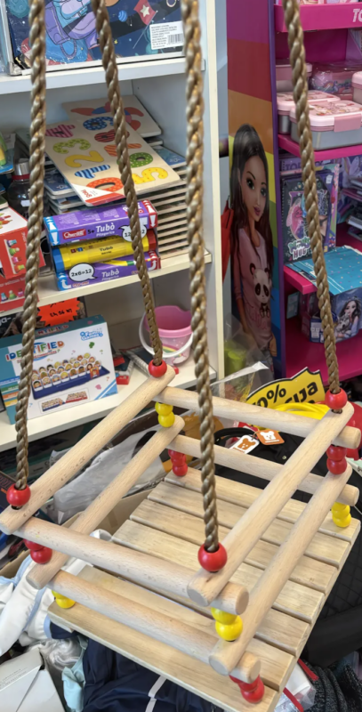 Suspended wooden toddler swing with rope and colored beads, displayed in a toy store aisle with puzzles and dolls nearby.